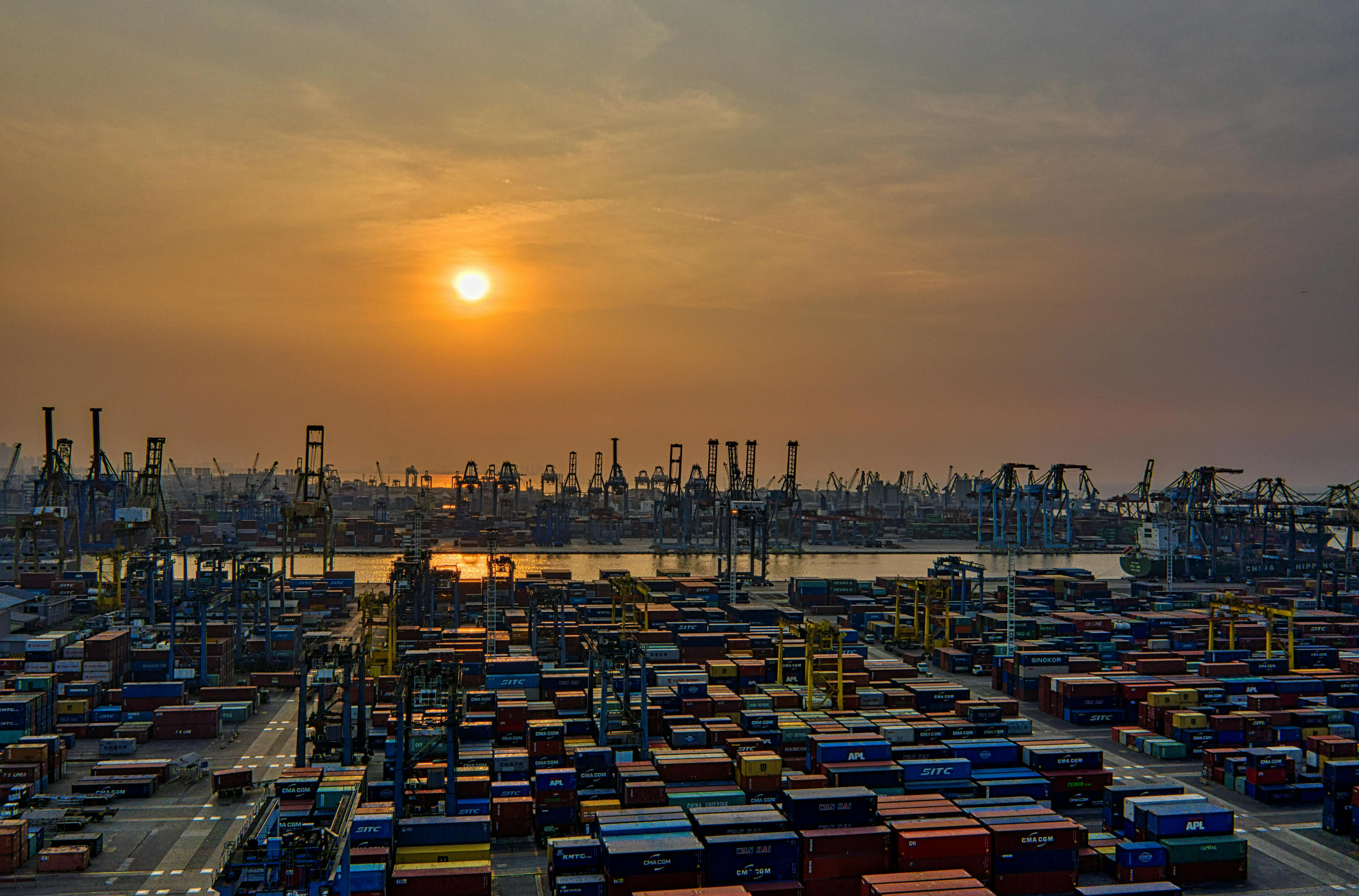 European container port fulled with containers, with cranes and the sunset in the background.