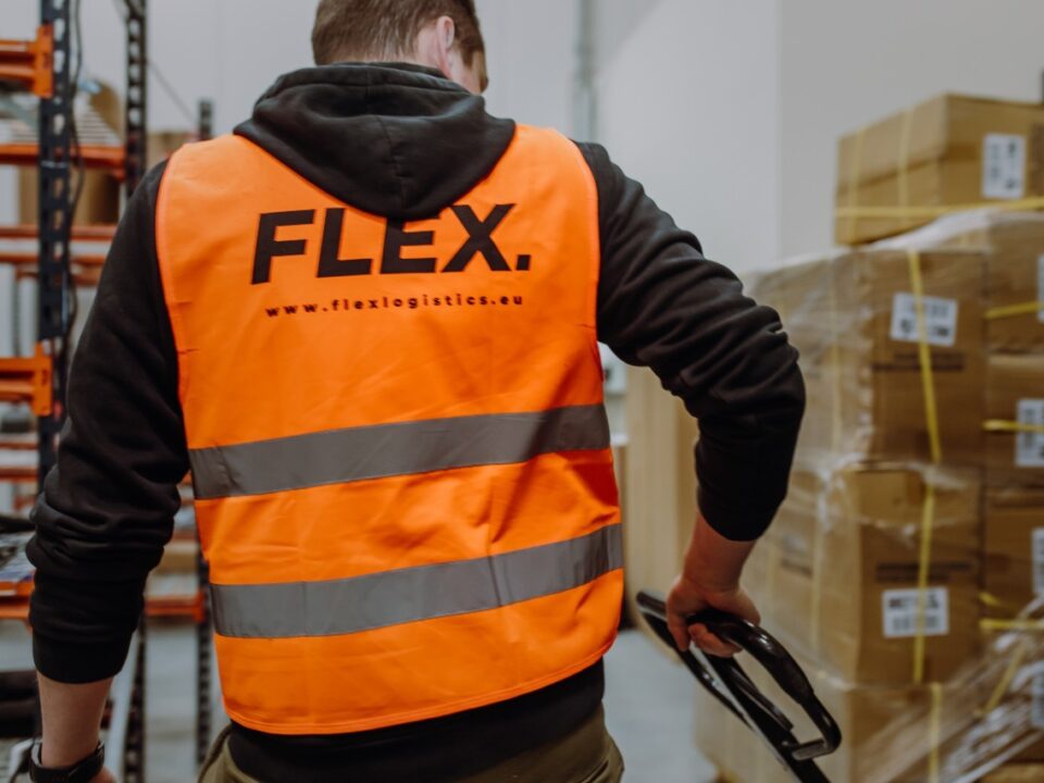 Worker in an orange "FLEX" safety vest operates a pallet jack in a warehouse filled with stacked boxes, suggesting efficient logistics work.