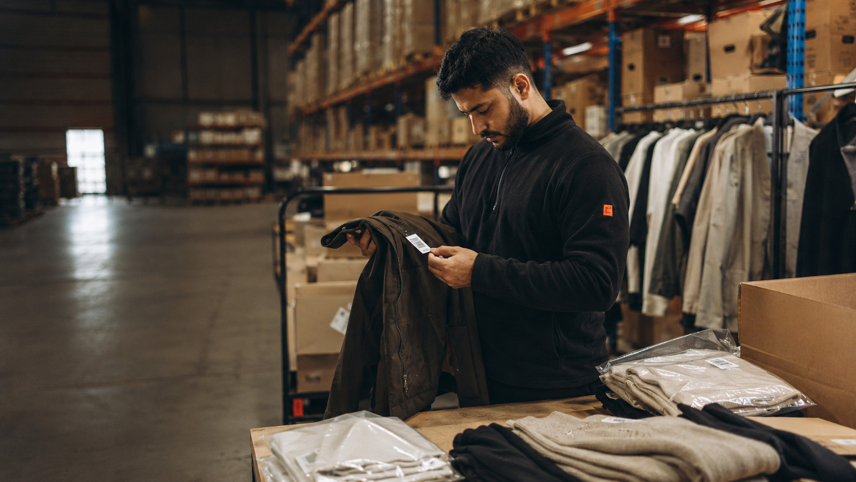 Apparel returns being inspected and sorted on a warehouse table with hanging rails, cartons, and barcode labels nearby.