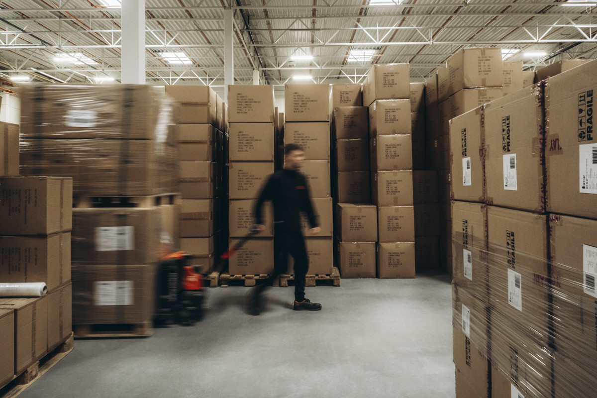Warehouse employee moving pallet handling equipment between large stacks of cartons and palletized inventory in a high-volume storage facility.