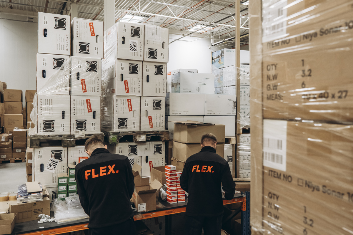 Two FLEX. warehouse employees processing and packing products at a fulfillment workstation surrounded by palletized inventory and boxed goods.